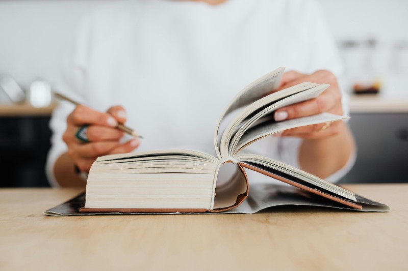 Людина перегортає сторінки товстої книги й робить нотатки ручкою на аркуші, сидячи за столом